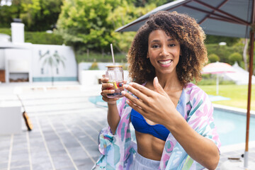 African American woman standing on pool deck holding fruit infused jar under umbrella beside guitar