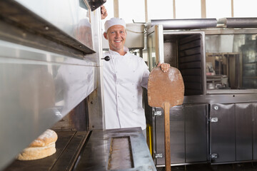 Mature male baker sliding loaf from industrial oven with wooden peel in commercial bakery kitchen