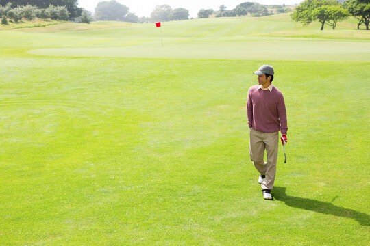 Naklejki Golfer walking across vibrant green fairway toward red flag, holding putter with red glove