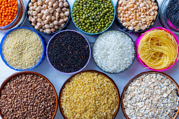 Assorted dry grains, legumes, and pasta in colorful bowls. A top view of various dry grains, legumes, and pasta types arranged in colorful bowls on a white background, including lentils, chickpeas