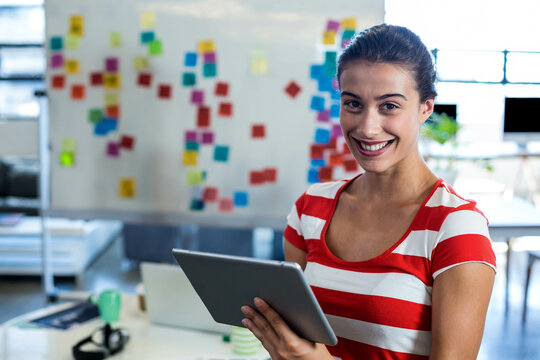 Mid-20s woman smiling and holding tablet in office by whiteboard with sticky notes, copy space