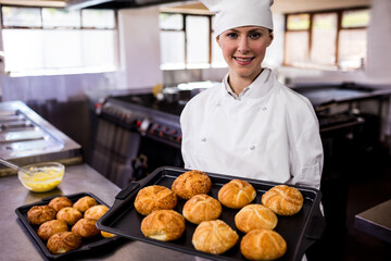 Female chef in chef coat and hat, holding baking tray with golden rolls in restaurant kitchen
