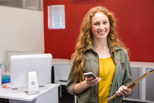Female intern wearing green jacket, holding smartphone and clipboard in computer lab with monitors