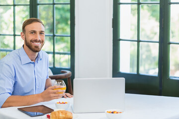Man sitting at dining table holding glass of orange juice with laptop, tablet, bowls, copy space © WavebreakMediaMicro
