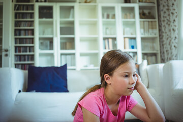 Preteen girl resting head on hand sitting by white sofa with cushion, bookshelf and floral curtains