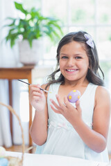 Naklejka premium School-age girl painting egg at table by bright window at home with wicker basket and paintbrush
