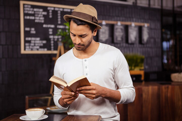 Bearded man reading paperback book by coffee cup on table in coffee shop with chalkboard menu