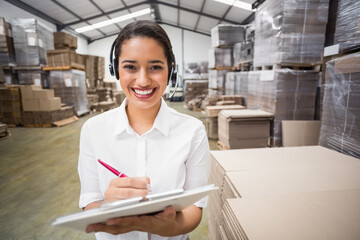African American woman inspecting boxes in warehouse holding clipboard, red pen and wearing headset