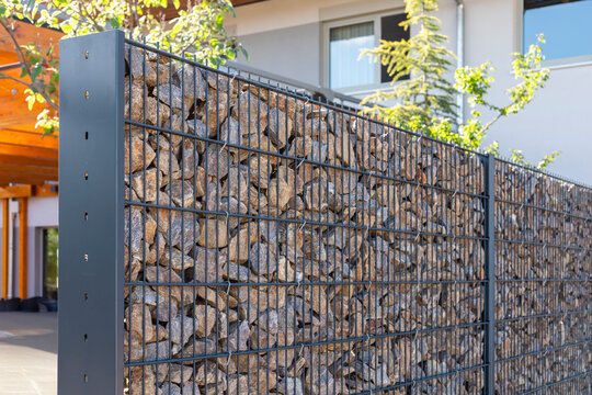 A modern gabion fence constructed from stones creates an elegant boundary. It stands in a contemporary setting, complemented by greenery and a sleek architectural backdrop, showcasing innovation