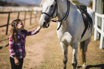 Girl standing beside light gray horse in ranch paddock petting muzzle near wooden rail fence
