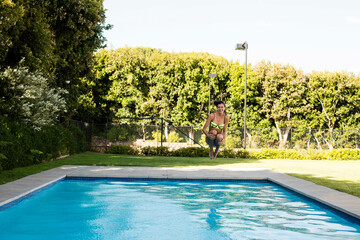 Female in 20s wearing neon green two-piece swimsuit doing tuck jump above backyard stone pool deck