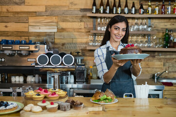 Female barista holding chocolate cake with raspberries at cafe counter with coffee tap, copy space