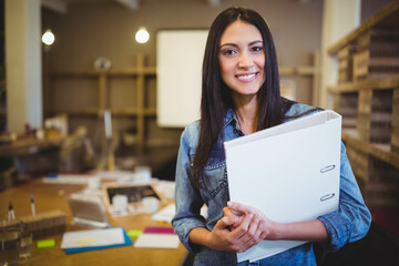 Fototapeta premium Woman holding white binder standing in studio, with wooden table covered in papers and markers