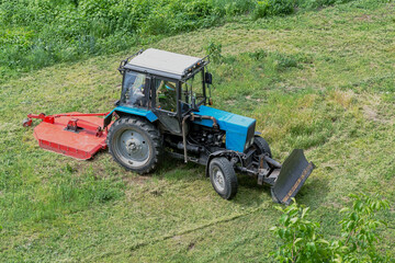 Obraz premium Tractor with a lawn mower during grass cutting, machine for grass cleaning, mowing grass and weeds in the field, tractor removing overgrown green grass.