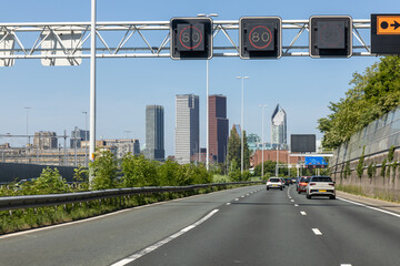 Urban highway A12 entering The Hague, Netherlands, with modern skyscrapers in the background and cars driving under digital speed limit signs.