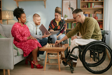 Diverse group of young adults and middle aged man with disability in wheelchair playing chess together in living room, Black woman and Caucasian man watching, multiethnic friends celebrating birthday