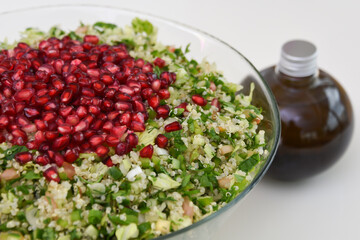 Fresh Tabbouleh salad in a clear glass bowl topped with pomegranate seeds, chopped parsley, bulgur, tomatoes, and mint with olive oil bottle closeup side view of healthy meal