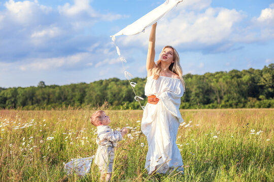 In a meadow in the grass and wildflowers, a child and a mother in a white dress and with blond hair are launching a kite into the sky, having fun with their little son.