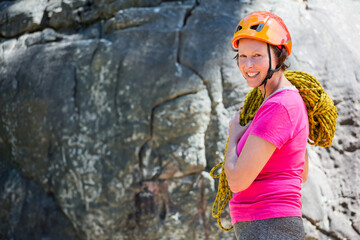 Woman standing at base of climbing cliff wearing orange helmet and carrying yellow rope, copy space