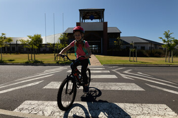 Girl wearing red helmet and teal backpack riding bicycle across zebra crosswalk at school entrance