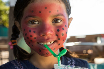 Girl with red-black-spotted face paint in shirt sipping frothy drink through straw at wooden table
