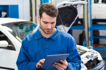 Male mechanic standing in workshop focusing on tablet with white compact car and lifted vehicle