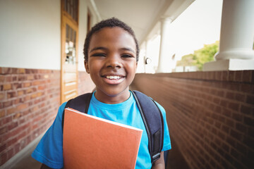Smiling boy in blue shirt carrying black backpack in brick corridor by door holding orange notebook
