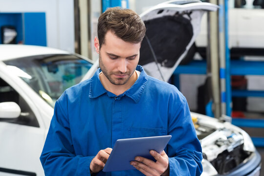 Male mechanic standing in workshop focusing on tablet with white compact car and lifted vehicle