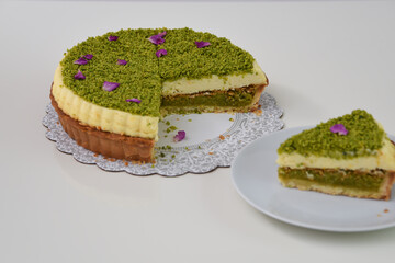 Delicious Pistachio Cheesecake with a slice removed presented on a white plate covered with finely ground pistachios and purple flower petals side view of elegant dessert