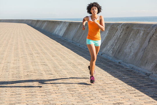 African American woman jogging along seaside path in orange tank running shoes beside sea barrier - Powered by Adobe