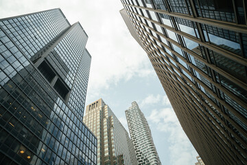An upward view of modern high-rise office buildings with glass facades and reflective windows. The photo captures a dramatic city perspective under a bright, slightly cloudy sky, showcasing urban