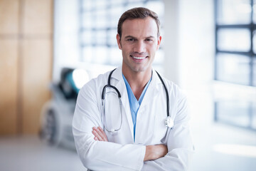 Doctor standing confidently crossing arms in medical corridor near windows, holding stethoscope