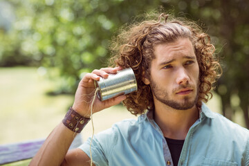 Adult man holding homemade tin can telephone and listening on bench in sunny green park