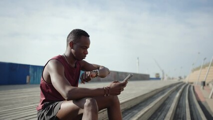 African American man sitting after training, checking fitness levels on smartwatch and using mobile phone app  - Powered by Adobe