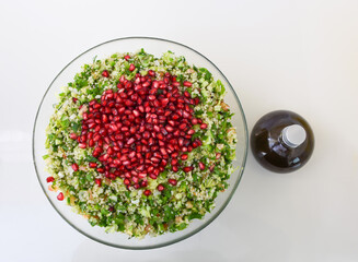 Fresh Tabbouleh salad in a clear glass bowl topped with pomegranate seeds, chopped parsley, bulgur, tomatoes, and mint with olive oil bottle top view of healthy meal