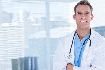 Male doctor standing by clinic window in scrubs and coat holding stethoscope by binders, copy space