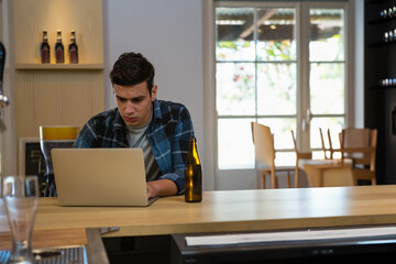 Man in his twenties working on laptop at home kitchen bar counter beside beer bottle