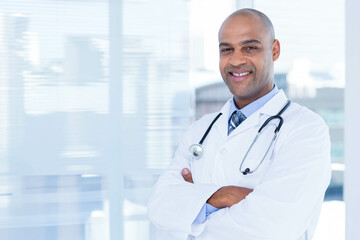 African American male doctor standing in clinic by blinds in lab coat, tie, stethoscope, copy space