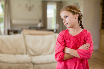 Naklejka premium Child crossing arms and wearing red top in room with beige sofa, wooden table and chandelier