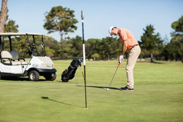 Golfer lining up putt on putting green near golf cart and bag under clear blue sky
