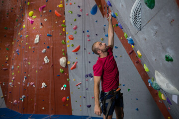 Climber scaling wall inside gym, using harness and rope with colorful holds on padded floor