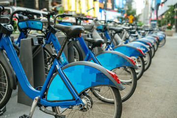 A row of blue public rental bikes is neatly lined up at a docking station on a busy city sidewalk. The background reveals blurred traffic, lights, and tall buildings, suggesting a vibrant urban