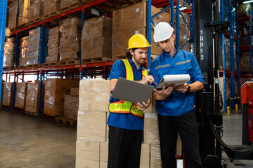 Two male warehouse workers in uniform and helmet standing beside stacked boxes and forklift,...