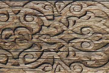 A close-up of a weathered wooden surface with intricate, ornamental carvings forming a symmetrical pattern. The aged wood and fine craftsmanship evoke traditional design and historical artistry.