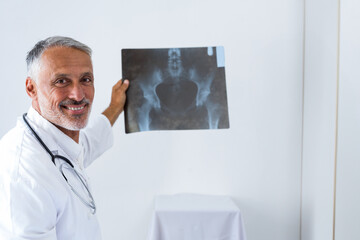 Middle-aged male doctor holding pelvic X-ray against wall in exam room with stethoscope, copy space
