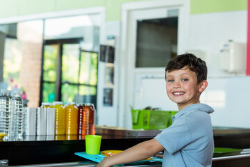 Smiling school-age boy holding lunch tray at school cafeteria counter by juice bottles, copy space © WavebreakMediaMicro