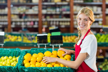 Smiling red-aproned woman arranging oranges in bins at grocery counter with price signs, copy space
