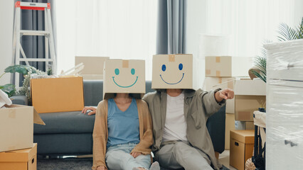 Happy Asian couple sit on sofa couch used carton box made smiling face funny dance front of camera in living room at new house. Husband and wife moving new house, Apartment Rent And Ownership Concept.