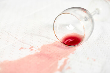 Red wine stain spreads across white textured fabric, highlighting contrast. Empty glass lies on side, causing spillage. Soft lighting enhances color intensity and texture details
