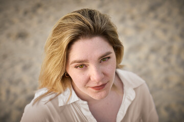 Young woman with blond hair gazes softly towards camera, standing on sandy beach. Her white shirt...